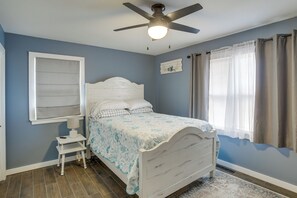 Un dormitorio con cama, ventilador de techo, ventana con cortinas y una mesita de noche.
