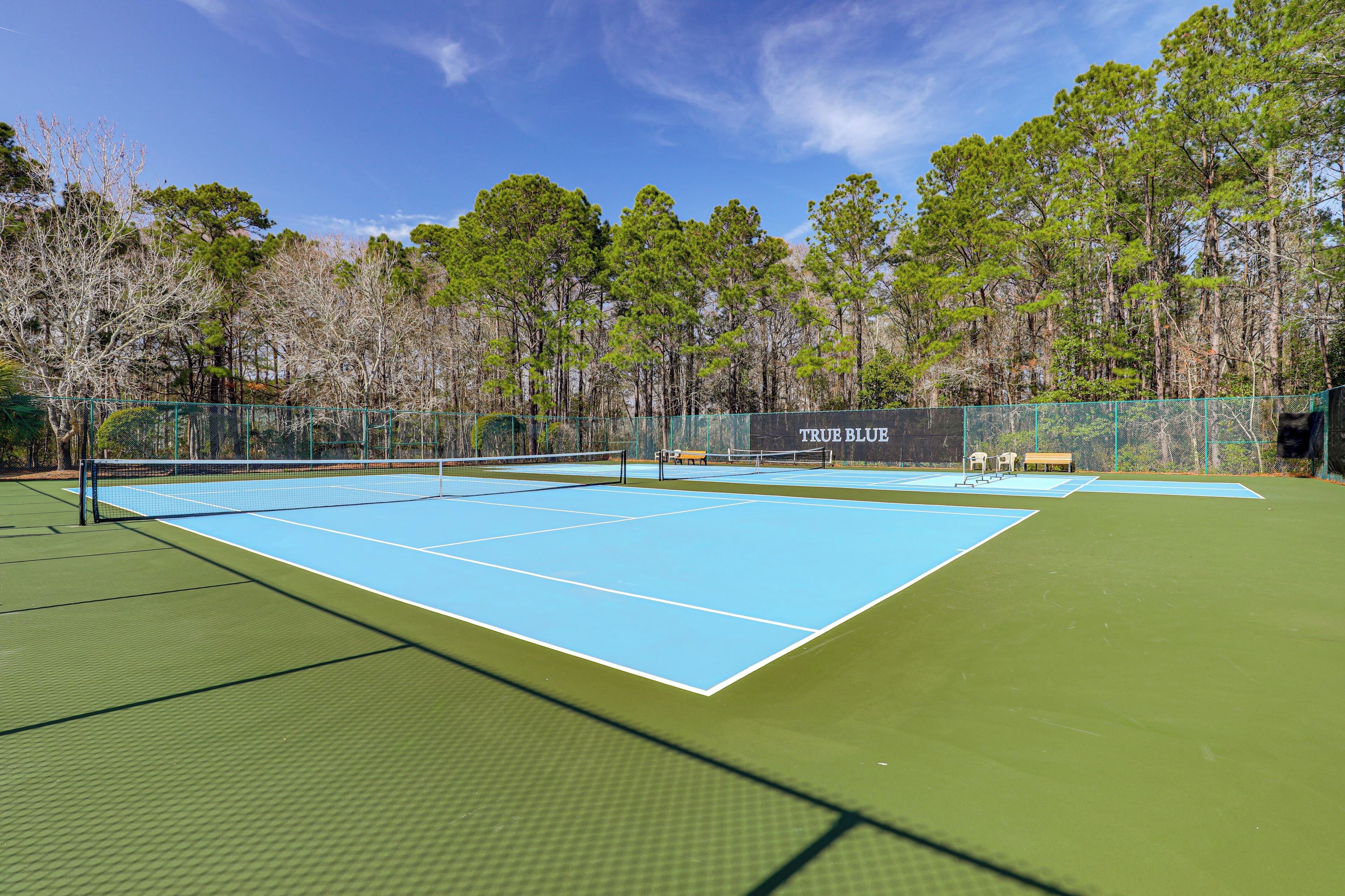Una cancha de tenis con red, rodeada por una cerca y árboles al fondo.