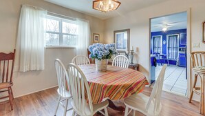 A dining area with a table, chairs, a chandelier, a window with curtains, and a view into a kitchen.