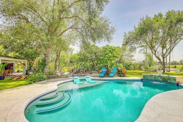 A swimming pool with a clear blue water feature, surrounded by lush greenery and outdoor seating. - Group Getaway: Luxe Riverfront Gem in Los Fresnos! (Los Fresnos)