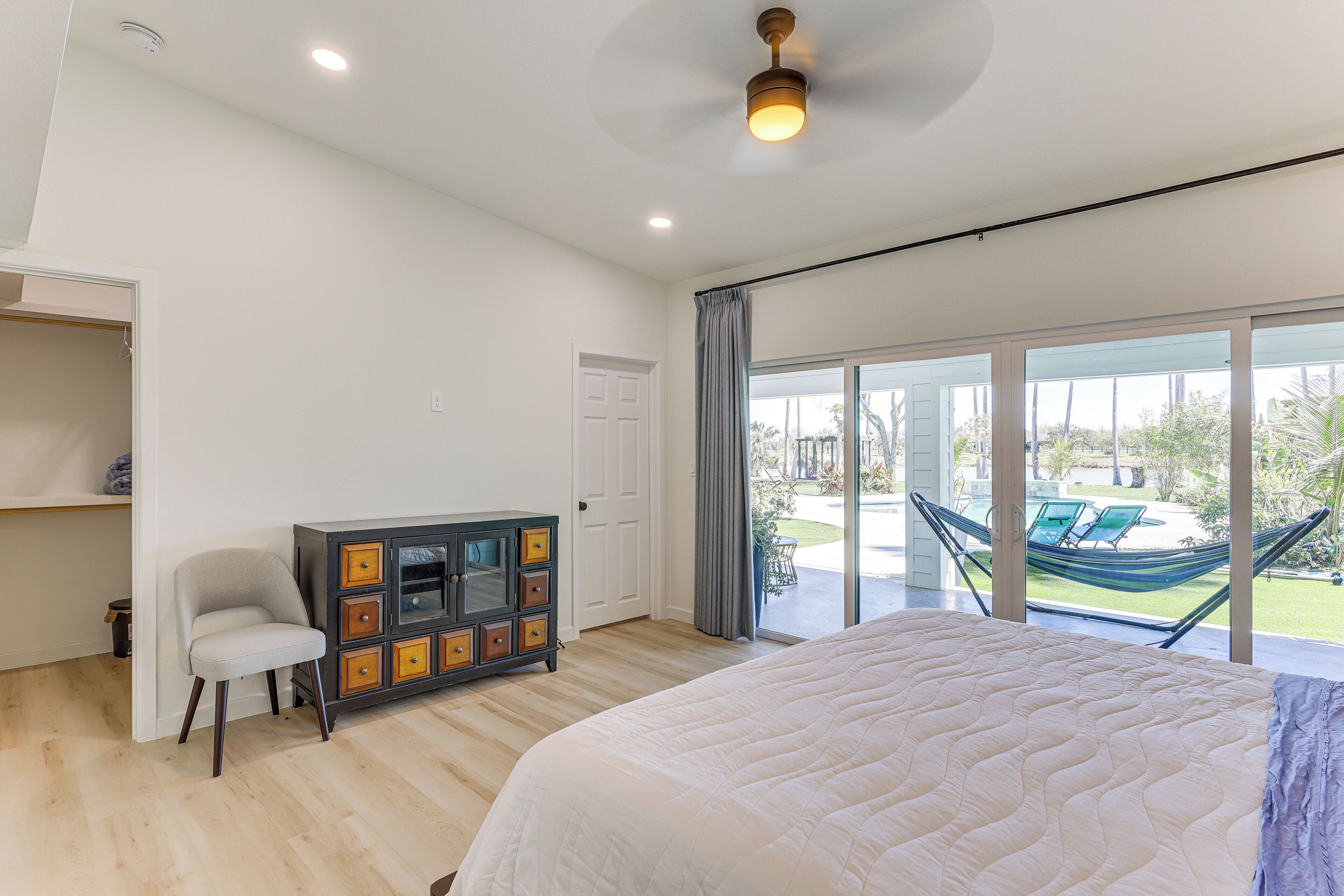 A bedroom with a large bed, a wardrobe, a chair, and a view of a hammock and outdoor seating.