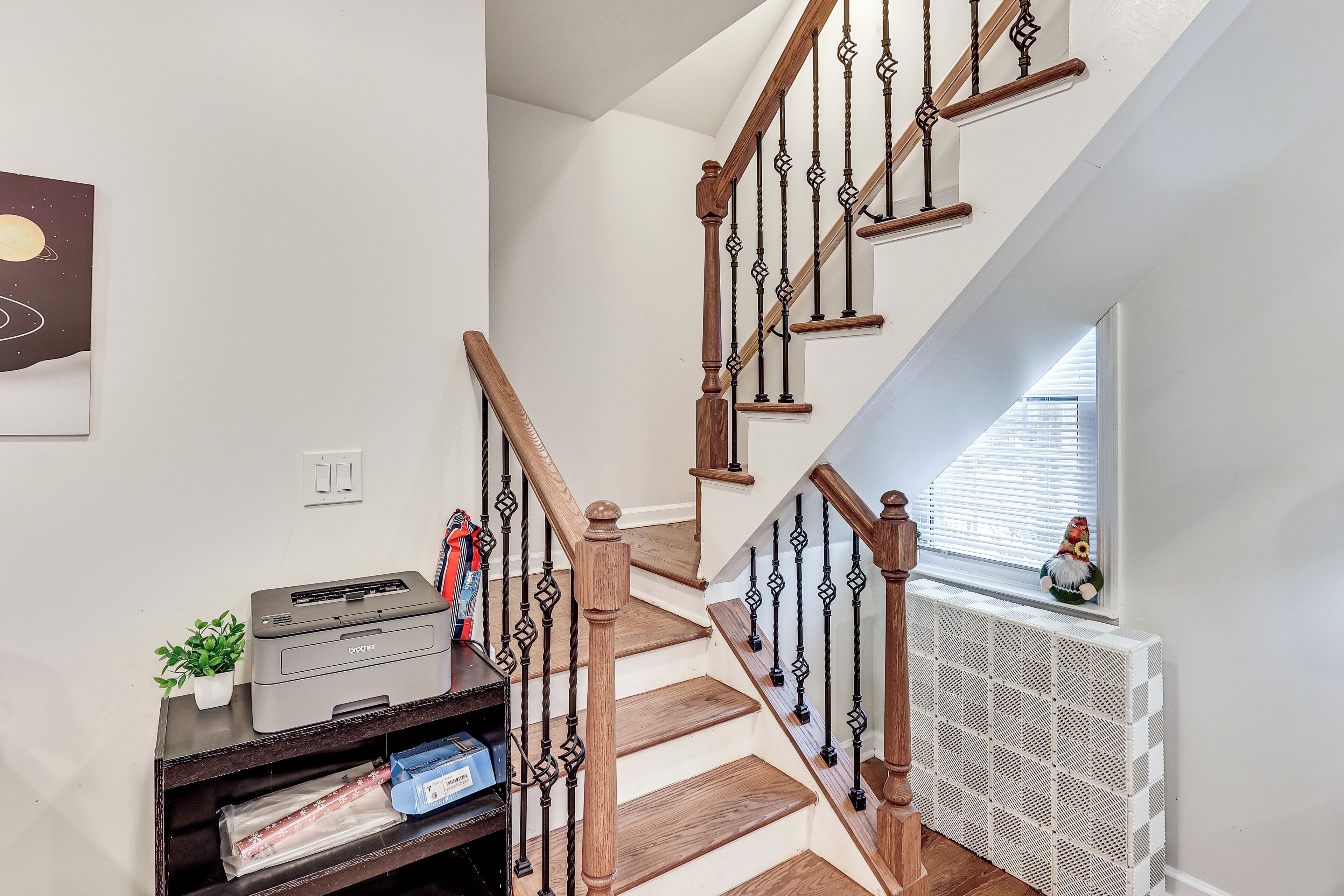Un escalier avec des marches en bois et des balustrades noires, un radiateur, une petite plante et une imprimante posée sur une étagère.