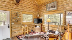 Ein Blockhaushaus-Interieur mit montiertem Tierkopf, einem Fernseher, einem Holzschrank, einer Ledersofa und einem großen Fenster.