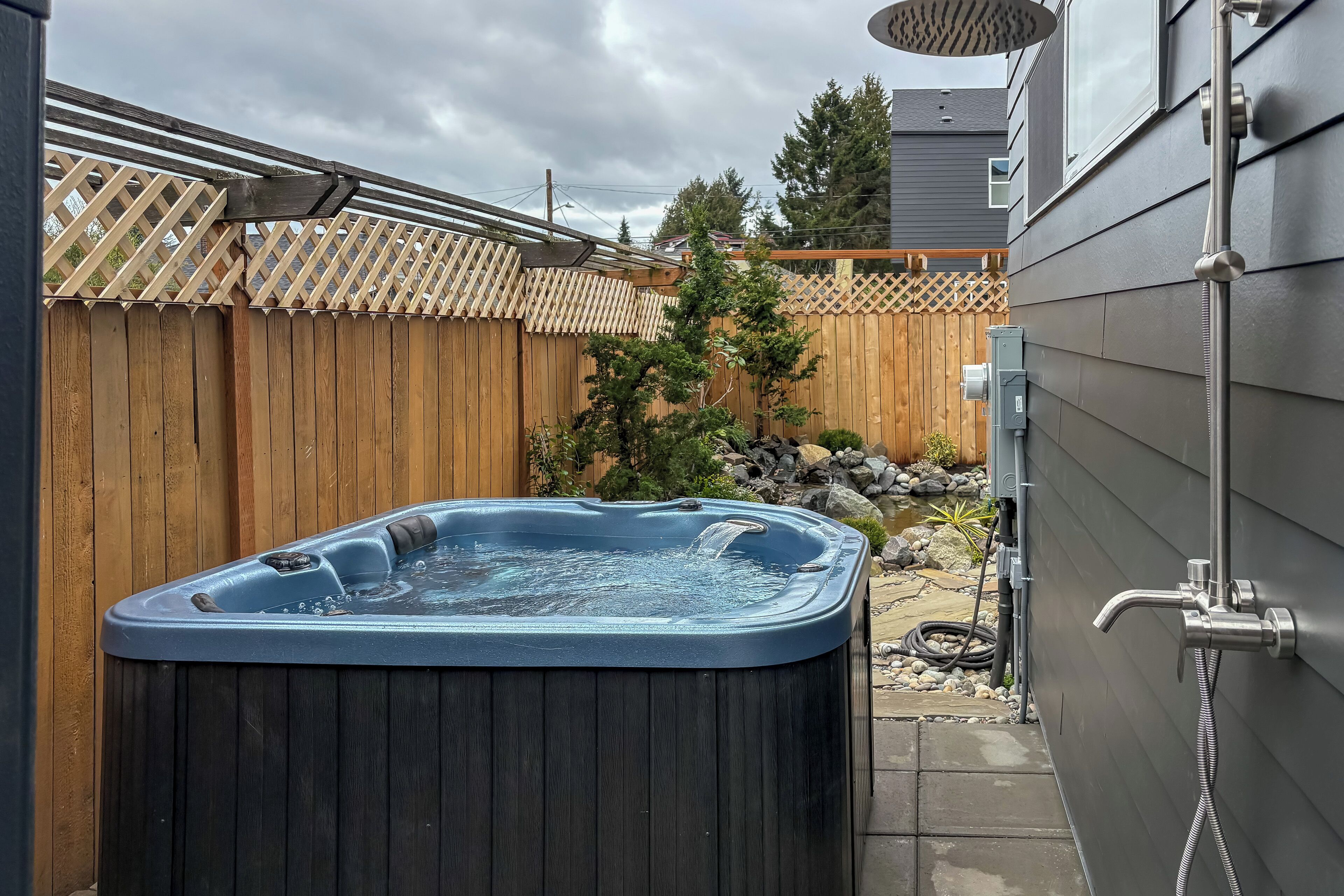 A hot tub in a backyard with a wooden fence, a small garden, and a metal showerhead.