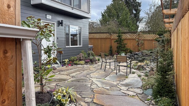 Une terrasse en pierre avec une clôture en bois, une terrasse en bois et un petit espace jardin agrémenté de diverses plantes et d’un banc.