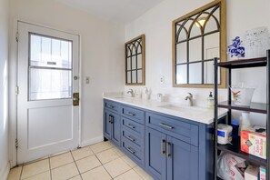 A bathroom with a white door, two sinks, a mirror, and a shelf with toiletries. - Centrally Located in Socal! Home Close to Highways (Bellflower)