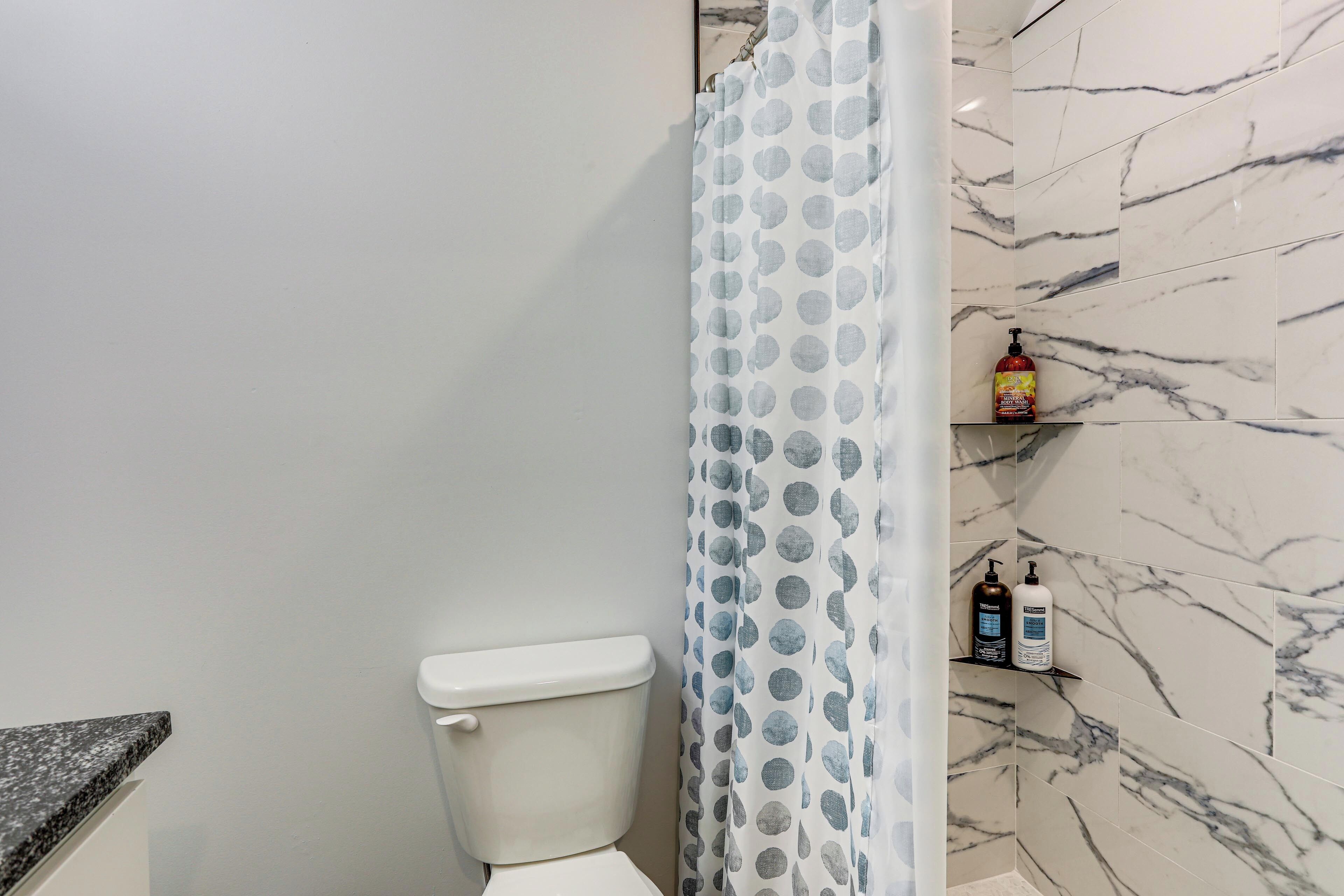 Une salle de bain avec des toilettes, un rideau de douche à motifs à pois et des murs à l’imitation du marbre. Il y a deux bouteilles sur une étagère.
