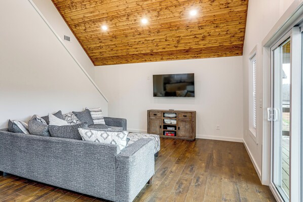 A modern living room with a wooden ceiling, a grey sofa, a flat-screen TV, and a wooden cabinet. - Ozarks Escape w/ River Views & Fire Pit (Van Buren)