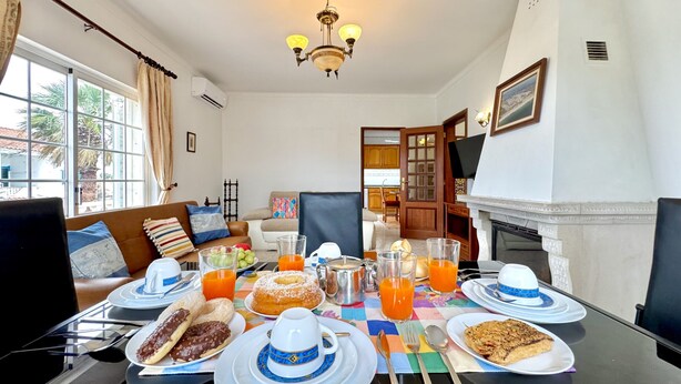Uma mesa de jantar posta com itens de café da manhã, um sofá e uma lareira em um ambiente bem iluminado.