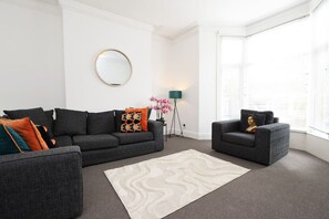 A living room with a dark grey sofa, a matching armchair, a patterned rug, a floor lamp, and a large window with blinds. - Spacious 6-Bed Home for Work Crews & Groups (Brynmill)
