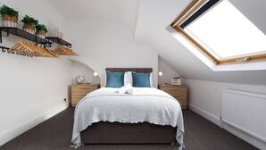 A bedroom with a skylight, a bed with a white bedspread, two nightstands with lamps, and a shelf with baskets and hangers.
