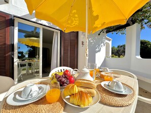 Un coin petit-déjeuner aménagé à l’extérieur, avec une table, un parasol jaune et divers éléments pour le petit-déjeuner.