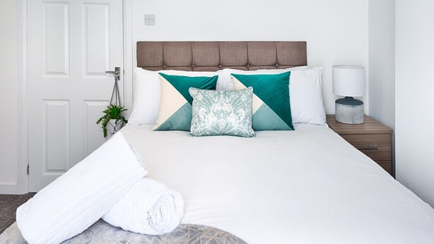 A neatly made bed with a headboard, a bedside lamp, and a rolled towel on the floor. - 4-Bed Swansea City Home | Walk to Centre | Ideal for Groups (Swansea)