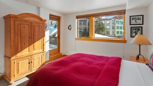 A bedroom with a large bed, a wooden wardrobe, a window with a view of buildings, and a lamp on a bedside table. - 4208 - One Bedroom Standard Creekside Condominiums 1 Condo (Solitude)