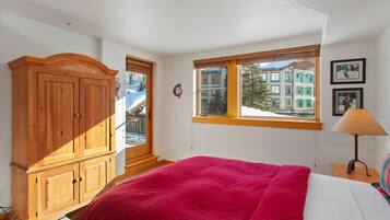 A bedroom with a large bed, a wooden wardrobe, a window with a view of buildings, and a lamp on a bedside table.