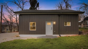 A single-story building with a gray exterior, a white door, and two windows with yellow lights. - Modern Arts District Loft With Boat Parking 1 Bedroom Cottage (Somerset)