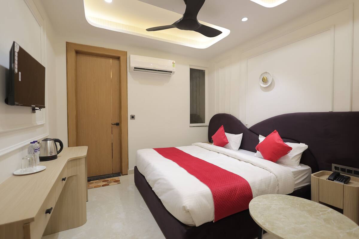 A hotel room with a bed, bedside table, a desk with a chair, a television, an air conditioning unit, and a ceiling fan.