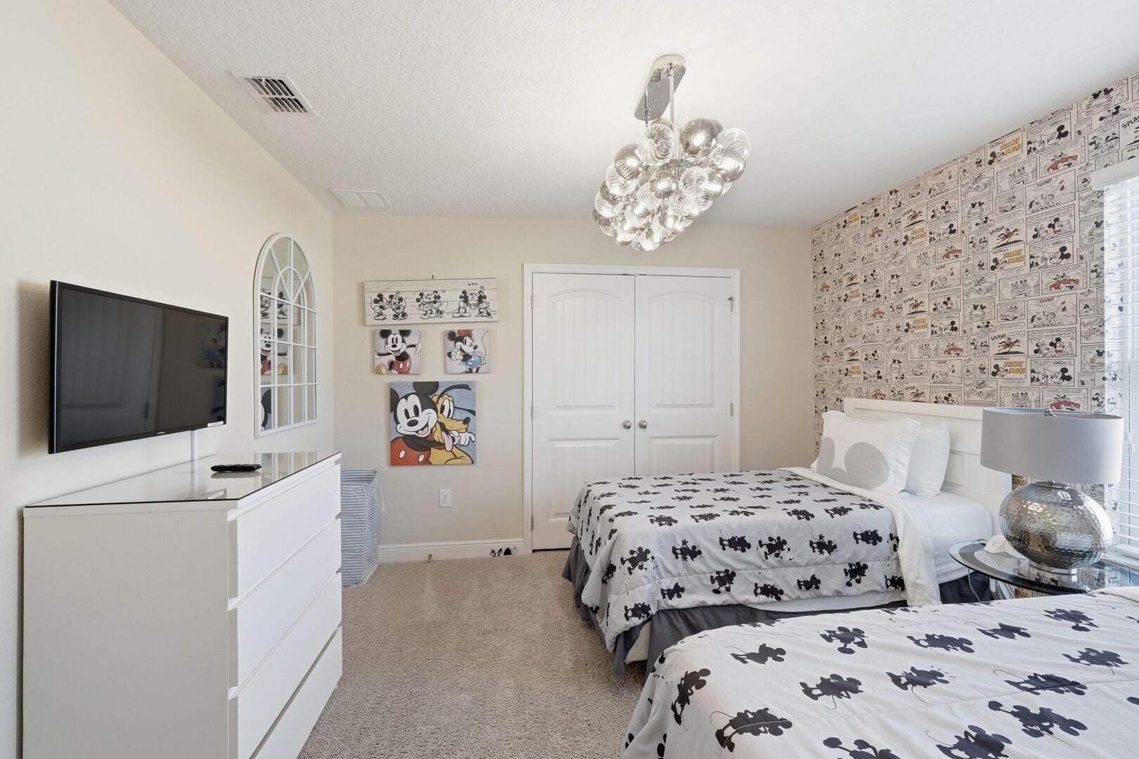 A bedroom with a television, a chandelier, a Mickey Mouse painting, and a Mickey Mouse themed wallpaper.