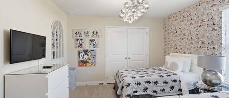 A bedroom with a television, a chandelier, a Mickey Mouse painting, and a Mickey Mouse themed wallpaper.