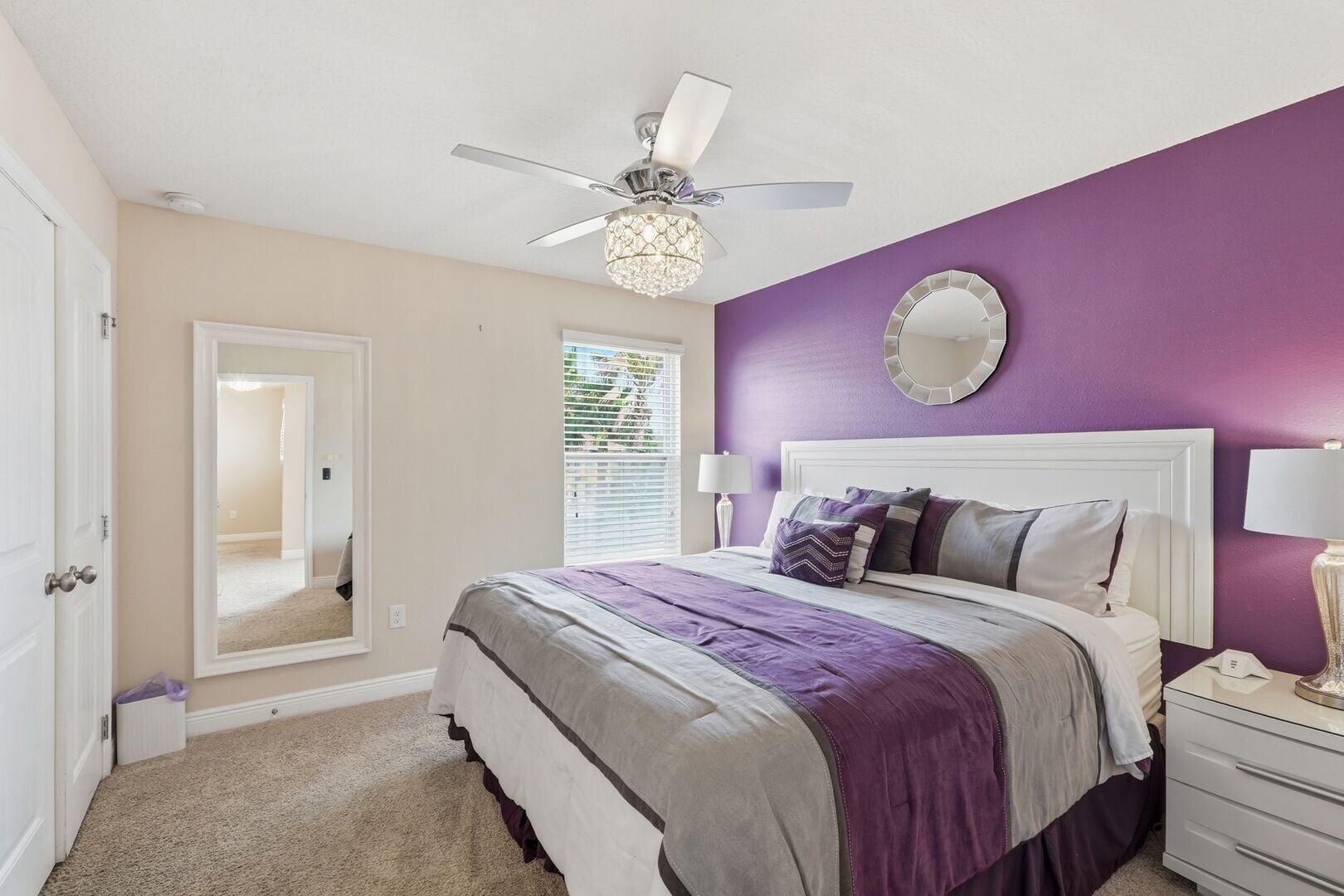 A bedroom with a bed, a ceiling fan, a window with blinds, a mirror, and a nightstand.