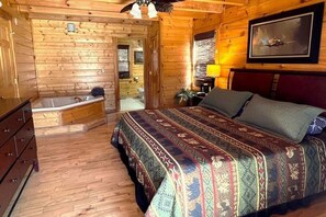 A log cabin-style room with a wooden floor, a large bed with a patterned bedspread, a wooden bench, a sink, and a window with a view. - Airport Runway 33 3 Bedroom Cabin (Pigeon Forge)