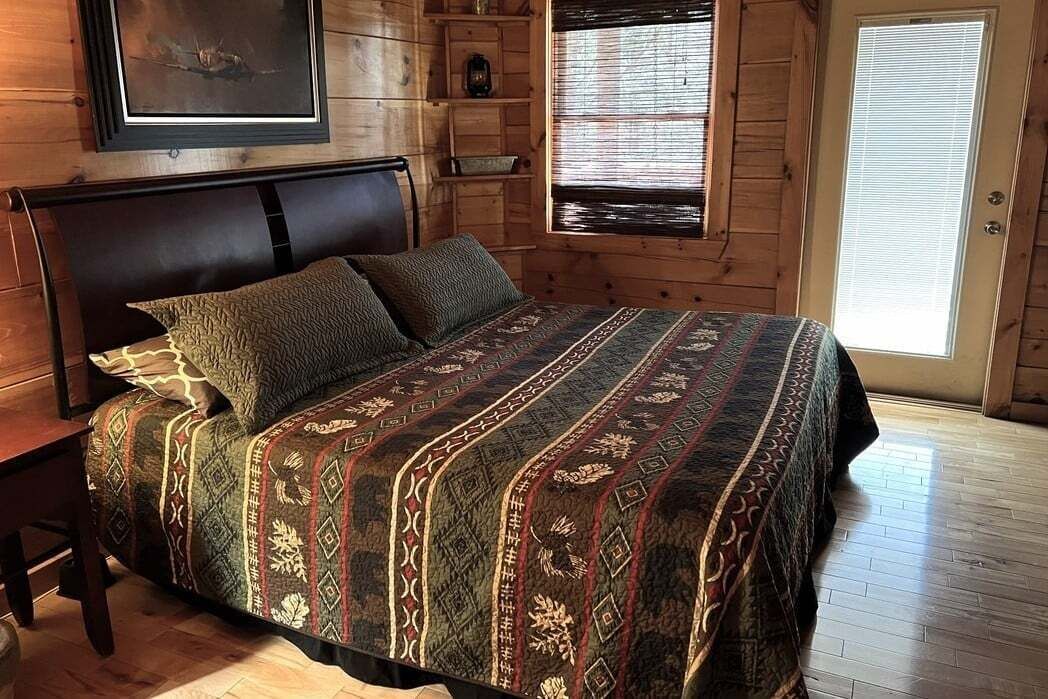 A wooden cabin-style bedroom with a large bed, a headboard, a window with blinds, and a door.