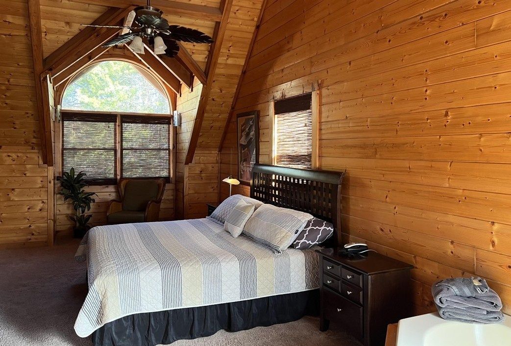Quarto com teto de madeira, cama com cabeceira, mesa de cabeceira, luminária e janela com persianas.