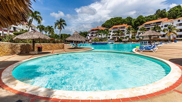 A large swimming pool with lounge chairs and umbrellas, surrounded by palm trees and hotel buildings. - Charming 2-bedroom Apartment in Sosua With Pool View (Sosúa)