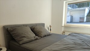 A bedroom with a bed, pillows, a nightstand, a lamp, and a window with a view of the outside.