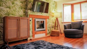Een woonkamer met een open haard, een aan de muur bevestigde televisie, een raam met zonwering en een ronde grijze fauteuil.