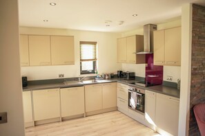 A kitchen with beige cabinets, a stainless steel range hood, a built-in oven, and a window with blinds. - Oak Lodge (Knighton)