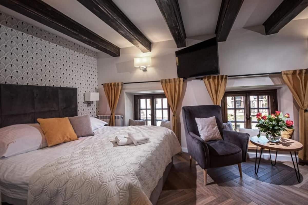Habitación de hotel con una cama grande, una silla, una pequeña mesa con flores y un televisor montado en la pared.