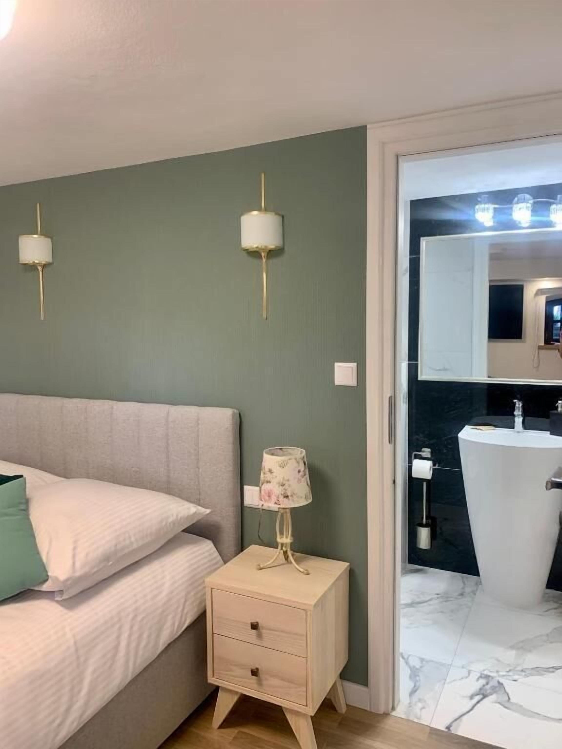 Una camera da letto con un letto, un comodino con una lampada, un punto luce a parete e un bagno con lavandino e specchio.