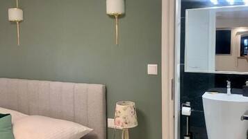 Una camera da letto con un letto, un comodino con una lampada, un punto luce a parete e un bagno con lavandino e specchio.