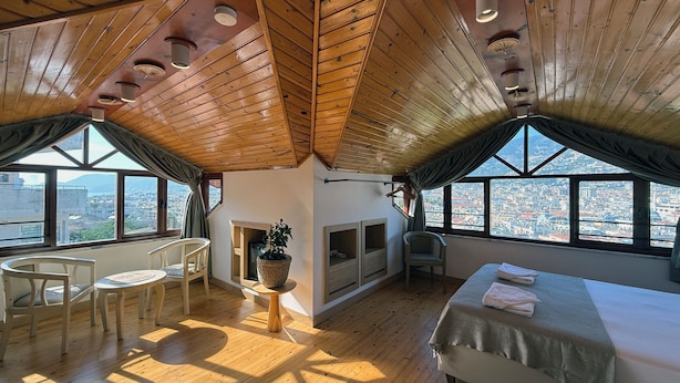 Una habitación con vigas de madera en el techo, una cama, una mesita con una planta, una silla y vistas de la ciudad a través de amplias ventanas.