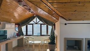 Una habitación acogedora con techo de madera, un ventanal con cortinas, una mesa redonda con sillas, una planta en maceta y un televisor sobre un escritorio de madera.