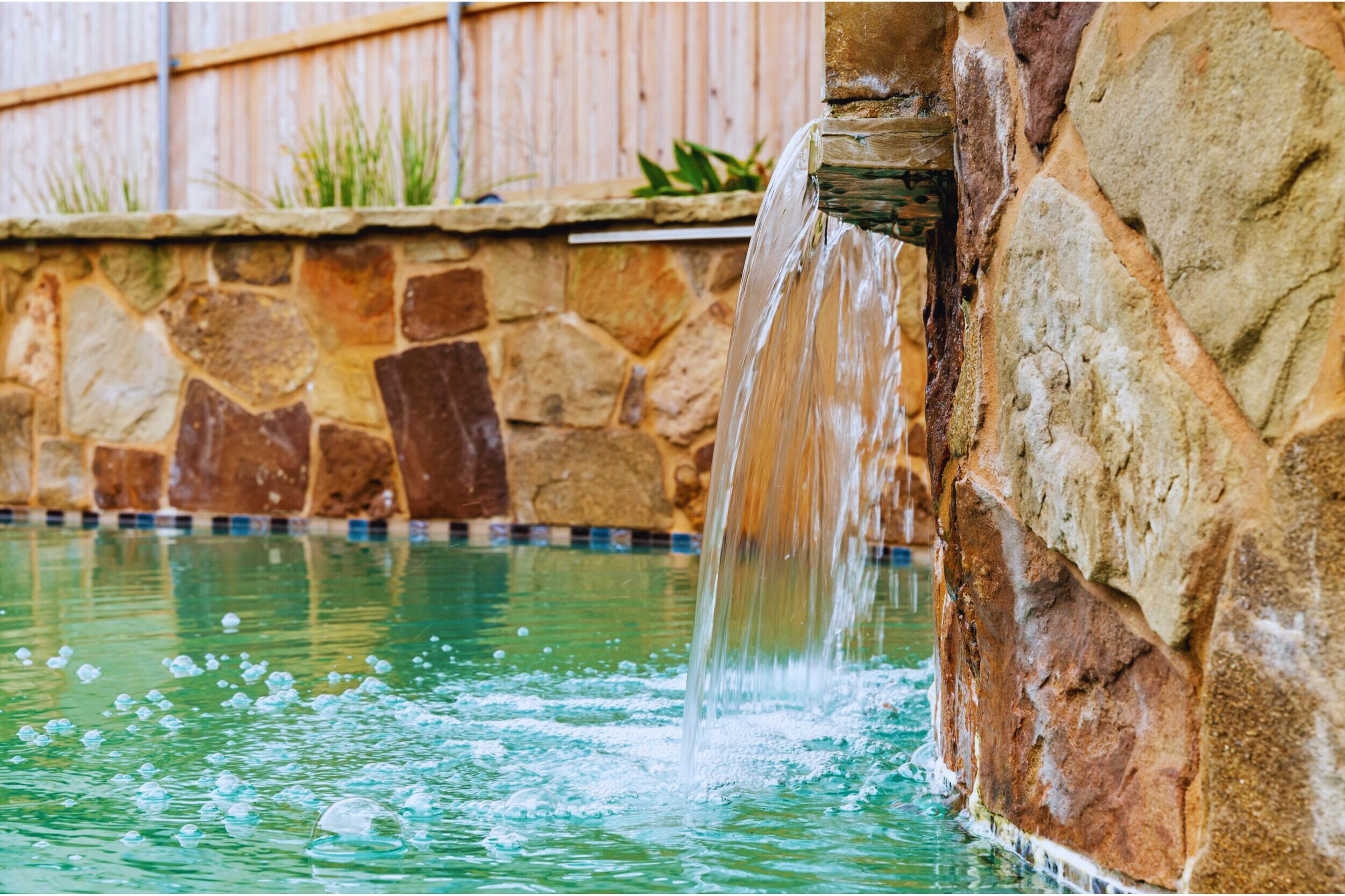 Eine Steinsäule mit Wasserspiel, Pool und Holzlattenzaun.