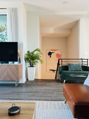 Ein modernes Wohnzimmer mit Flachbildfernseher, einem grünen Sofa, einem Couchtisch, einer Topfpflanze und einem Gemälde an der Wand.