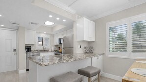 A modern kitchen with a marble countertop, stainless steel appliances, and a window with blinds. - Seaside 101 | Corner Condo Gulf View + Amenities (Fort Myers Beach)