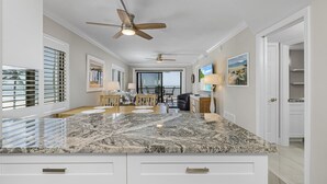 A kitchen with a granite countertop, white cabinetry, ceiling fans, and a dining area with a table and chairs. - Seaside 101 | Corner Condo Gulf View + Amenities (Fort Myers Beach)
