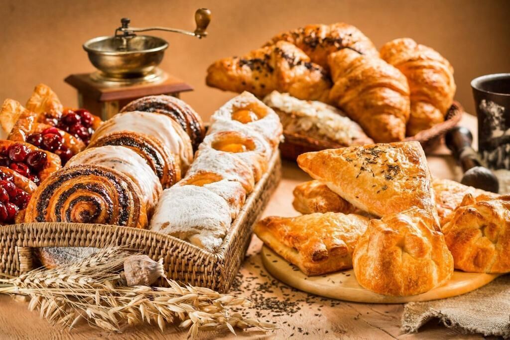 Variedad de pasteles y pan, incluyendo croissants y una cesta de pastelería con espigas de trigo.
