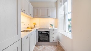 A compact kitchen with white cabinets, a built-in oven, a sink, and a window providing a view of the outside. - Modern Comfort in a Classic Bath Setting - VAT Q (Bath)