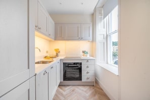 A compact kitchen with white cabinets, a built-in oven, a sink, and a window providing a view of the outside. - Modern Comfort in a Classic Bath Setting - VAT Q (Bath)