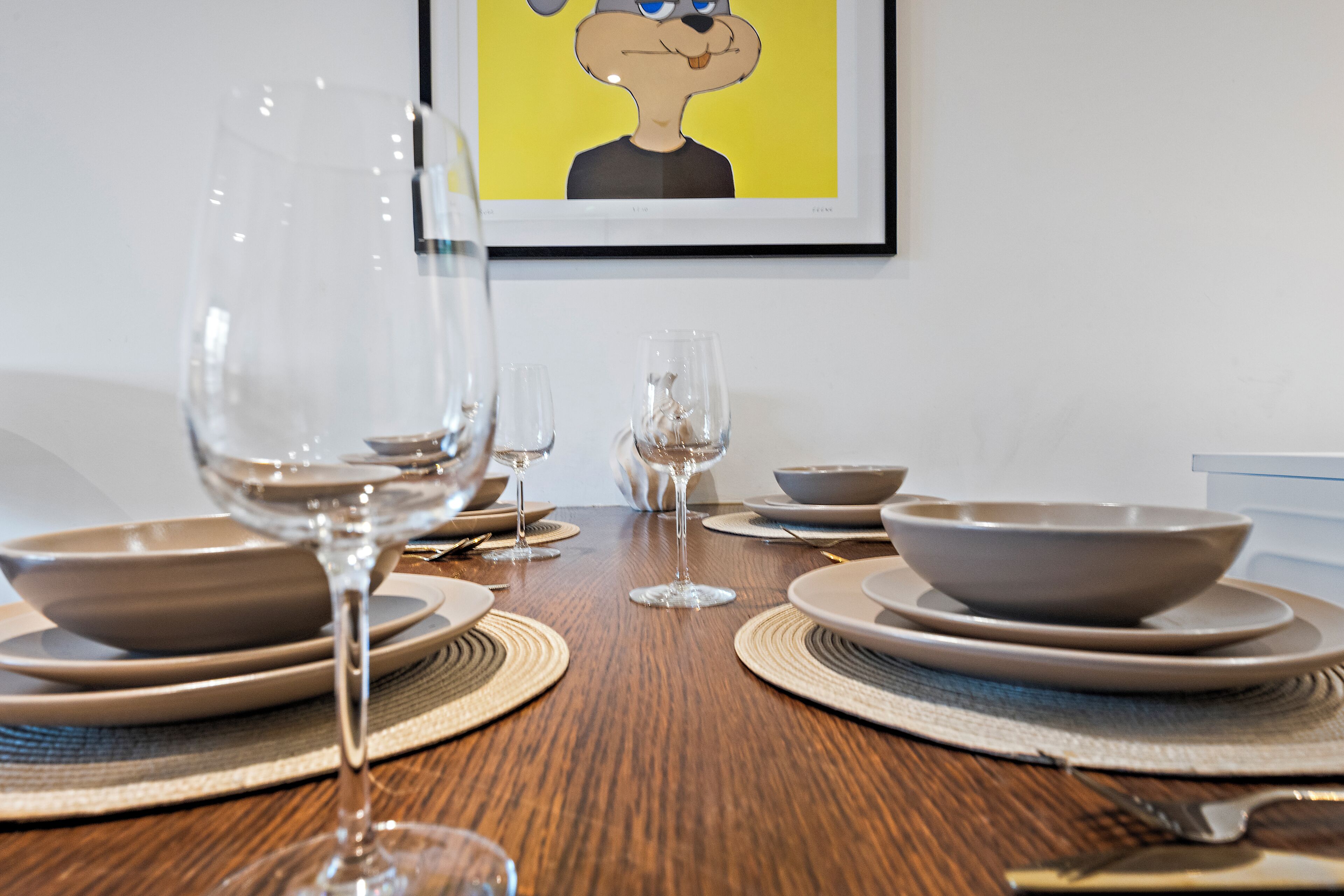 A dining table set with plates, bowls, and wine glasses, with a framed cartoon character on the wall behind.
