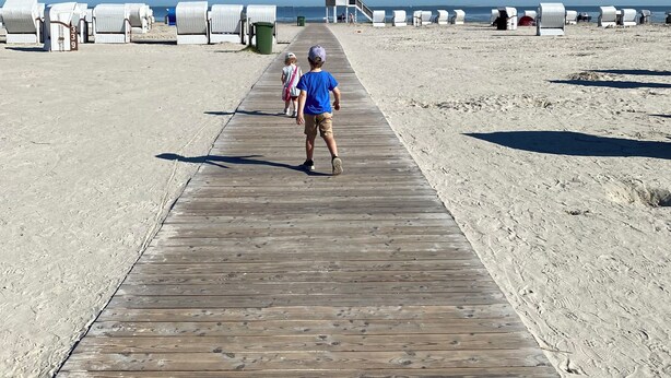 Une promenade en bois mène à des cabanes de plage, et on aperçoit une tour de surveillance des sauveteurs au loin.