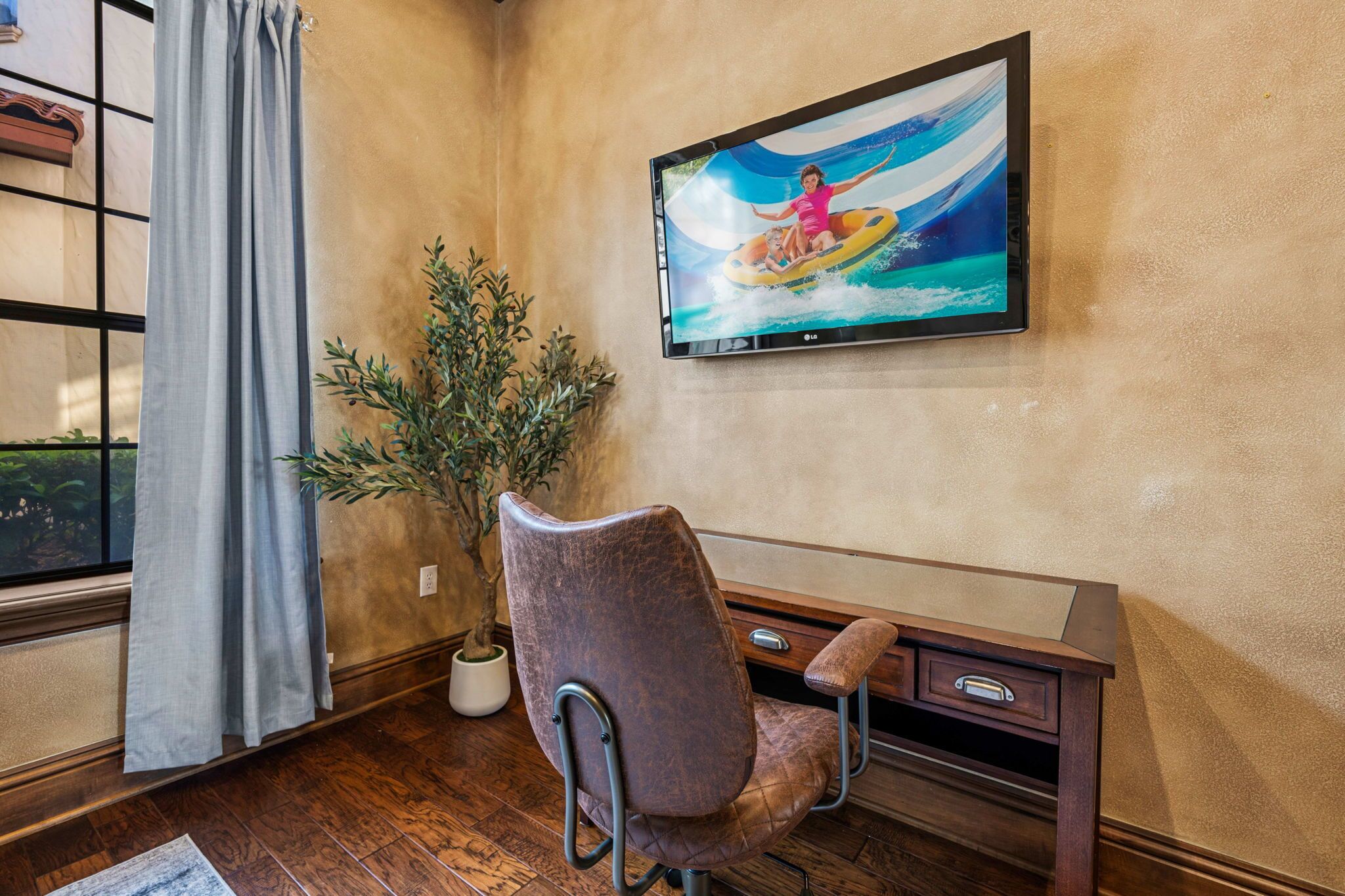 A room with a television mounted on the wall, a potted plant, a chair, and a wooden desk.