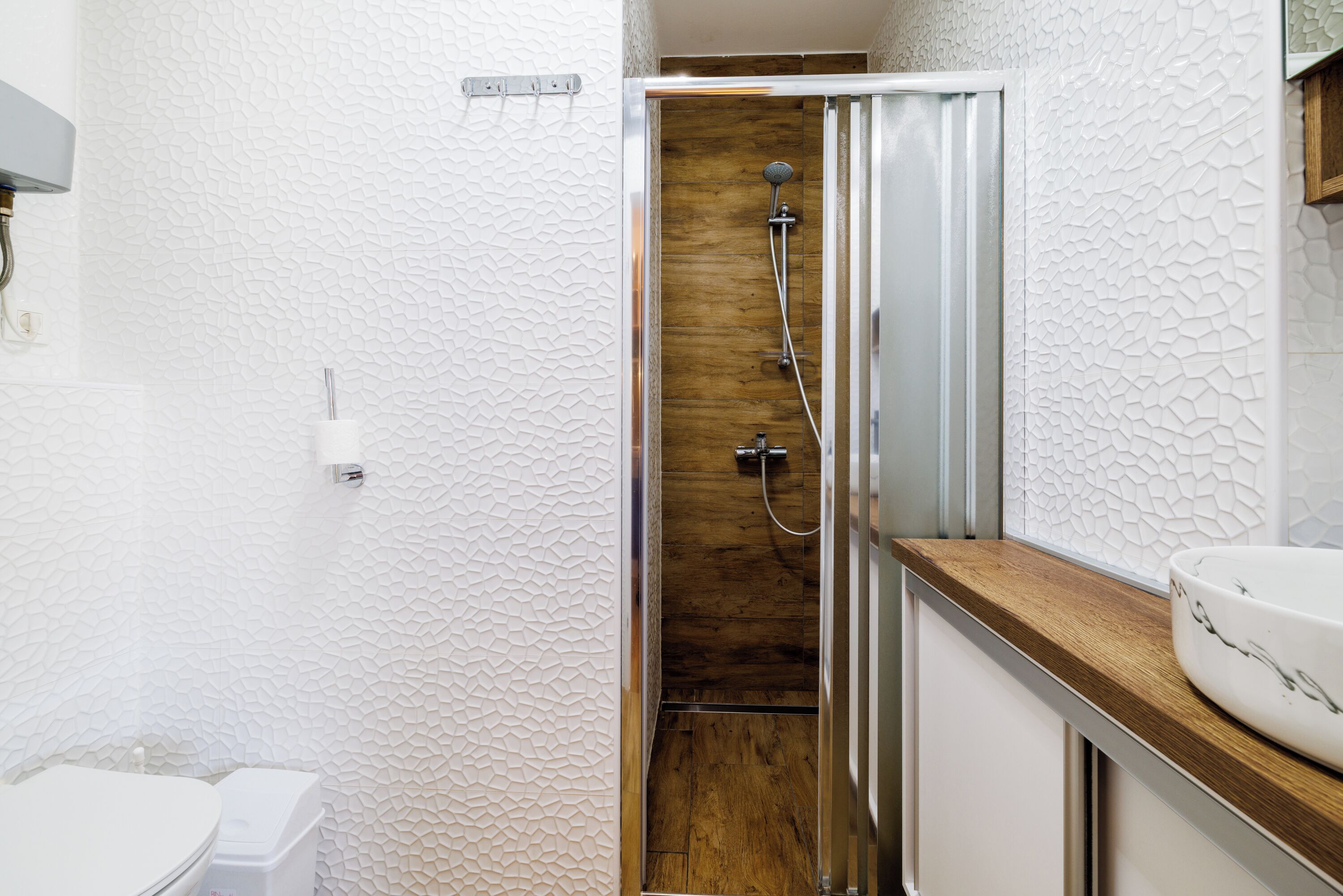 Un baño con pared blanca con textura, cabina de ducha de madera, inodoro blanco y encimera de madera con lavamanos.