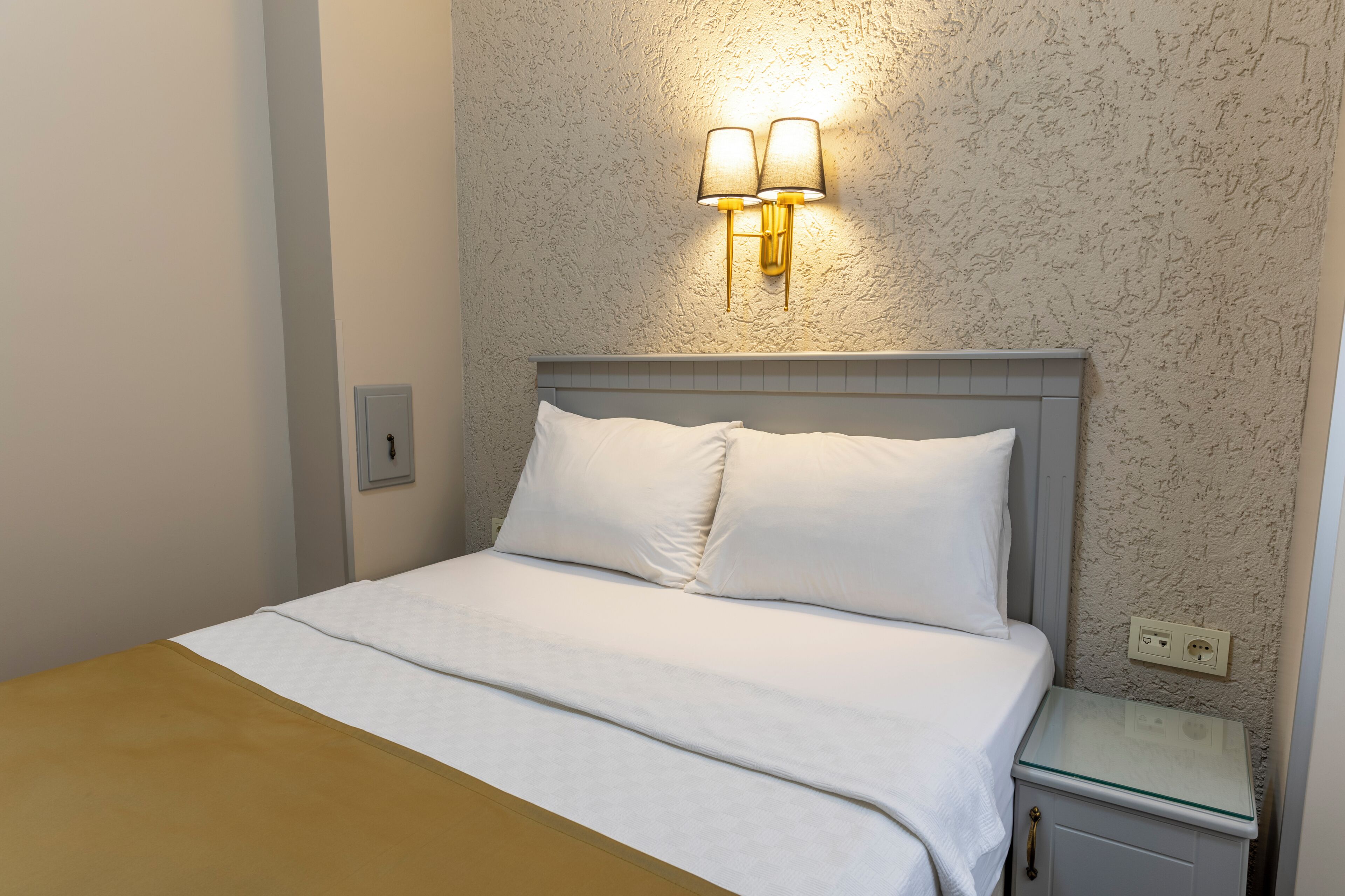 Una cama bien hecha con ropa blanca, una mesita de noche con superficie de vidrio y un aplique de pared.
