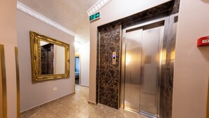 A hallway with a marble floor, a large mirror, an elevator, and a sign indicating the direction to the exit. - Hotel Umit 2 (İstanbul)