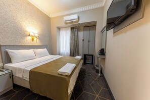 Et hotelværelse med seng, sengebord, lampe, aircondition, fjernsyn og et lille tekøkken.